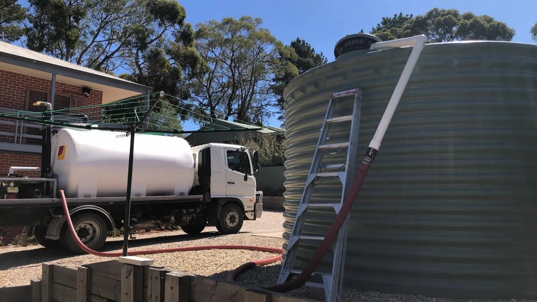 A water truck beside a large corrugated rainwater tank, its delivery hose run up a ladder into the top of the tank