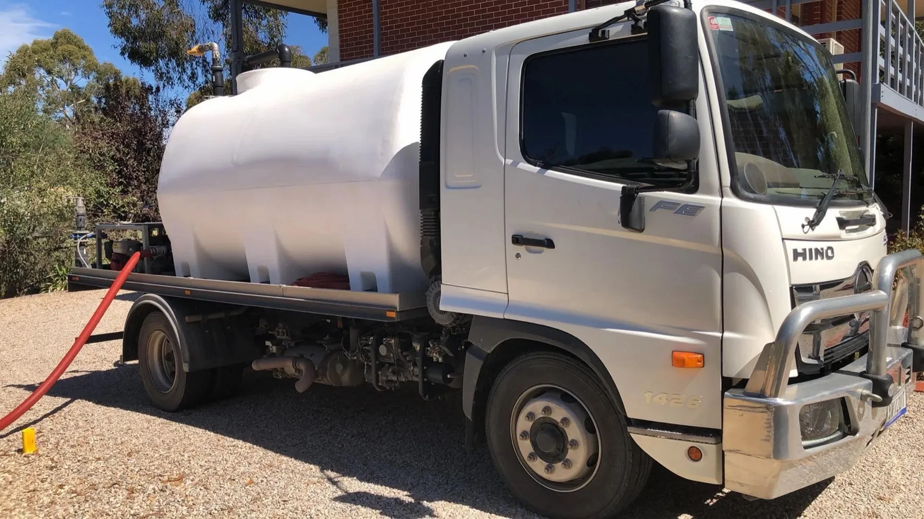 A white Hino water truck on a gravel driveway beside a brick house, its red delivery hose run out